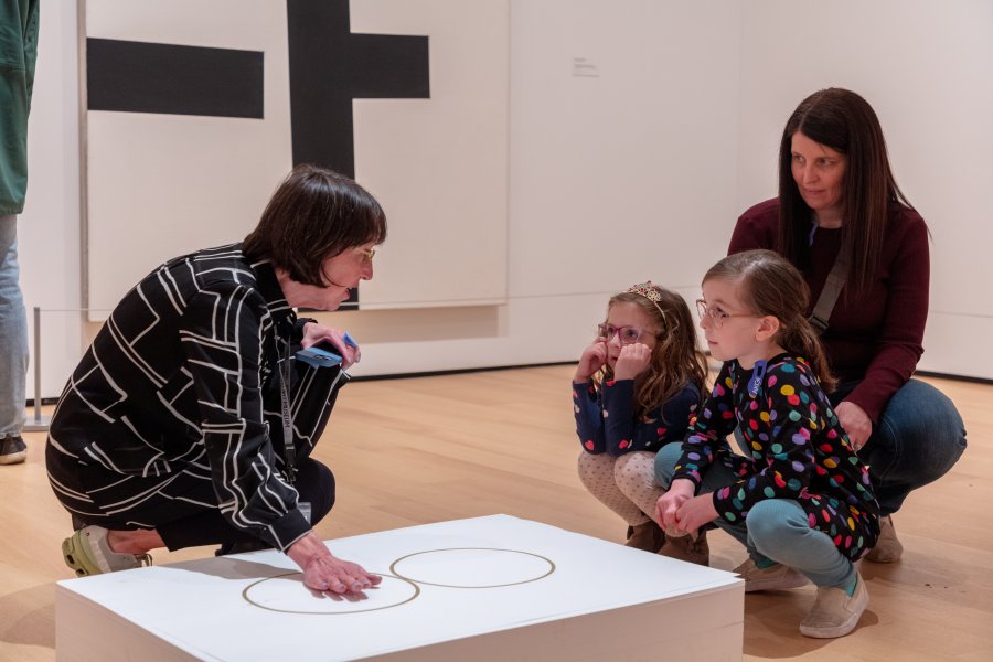 A docent explaining an artwork on the ground to two young girls and their mother